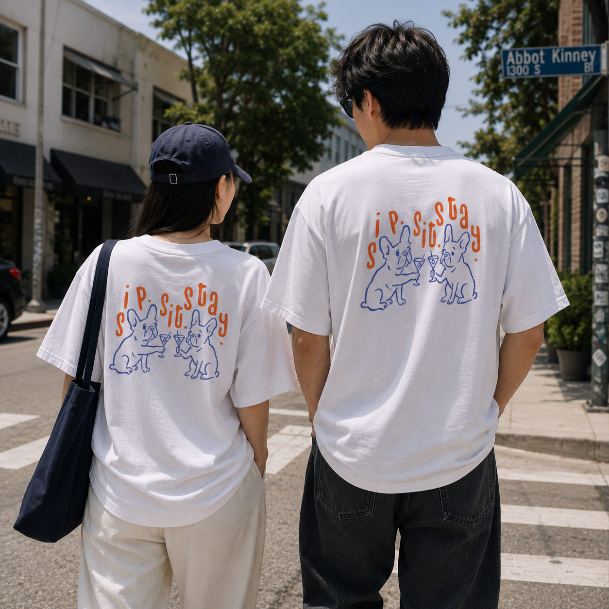 Two people standing on a city crosswalk, viewed from behind, wearing matching white graphic T-shirts with orange and blue cartoon dog designs and the text sipsitstay.