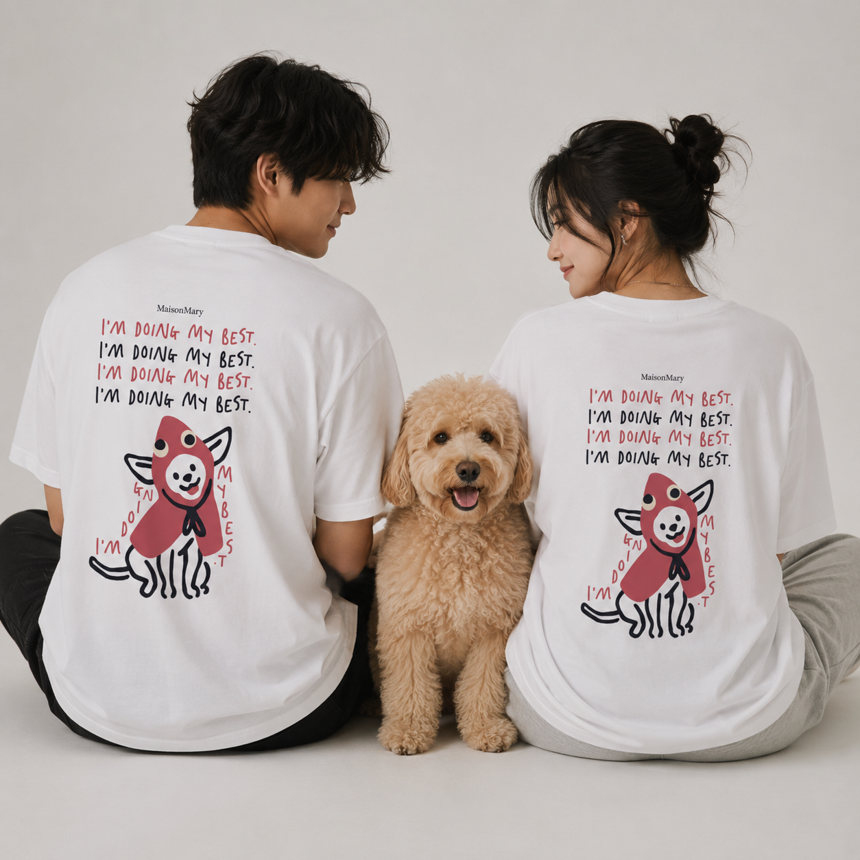 A man and a woman sit side by side facing away from the camera, wearing matching white graphic T-shirts with a red and black cartoon dog design and the text 'I'M DOING MY BEST.' A small tan dog sits between them on a light gray studio background.