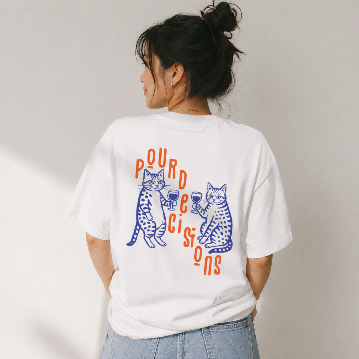 A woman shown from the back wearing a white graphic T-shirt with two blue cats and orange text reading 'pourdecisions' on the back, paired with light blue jeans against a plain light gray wall.