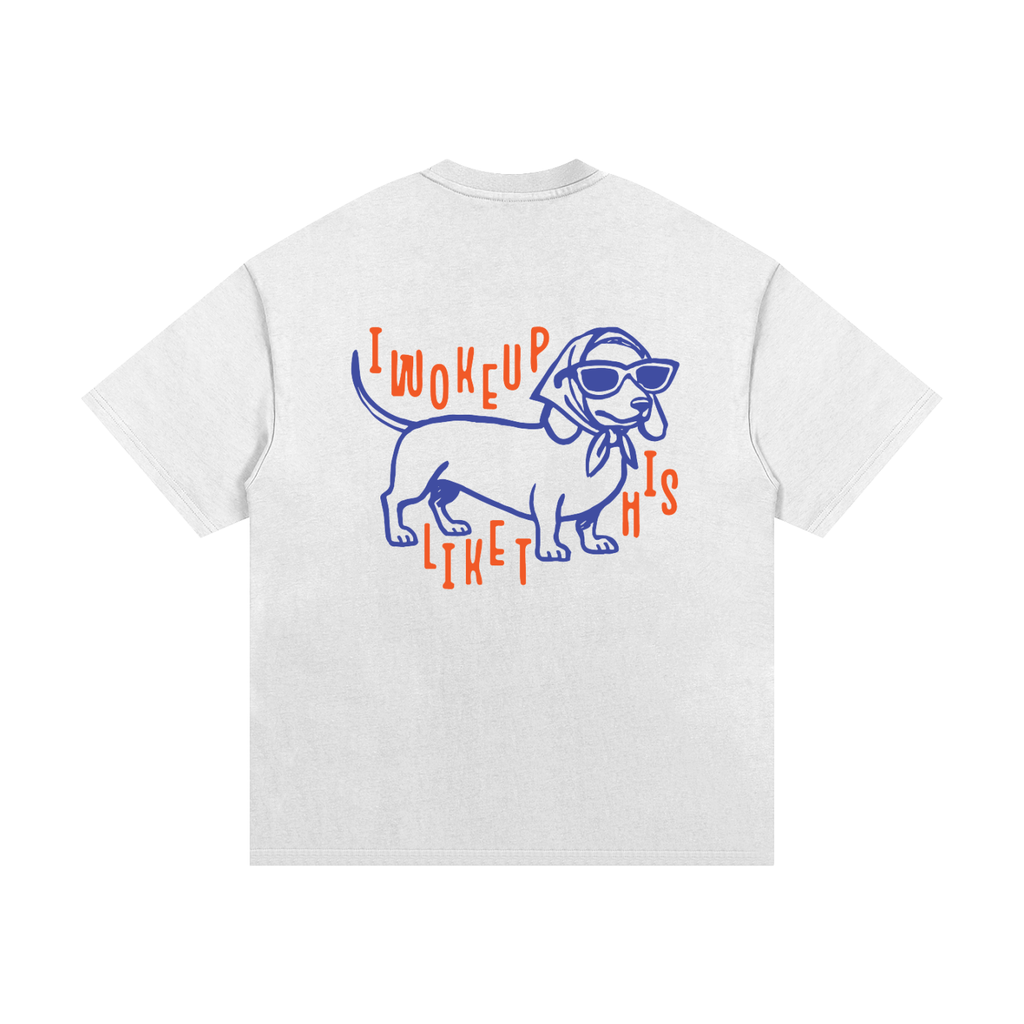 A white short-sleeve t-shirt shown from the back, featuring a graphic of a dachshund wearing blue sunglasses and a blue bow tie, surrounded by orange text that reads 'I WOKE UP LIKED THIS'.