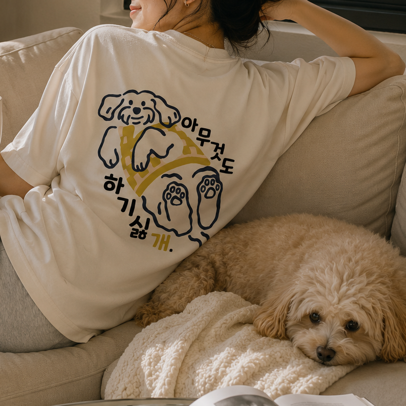 A woman relaxes on a beige sofa wearing a cream-colored pajama set with a graphic print of two dogs and Korean text. A small fluffy dog rests on a blanket next to her on the couch, and a mug with the text 'Home Sweet Home' is held in her hand.
