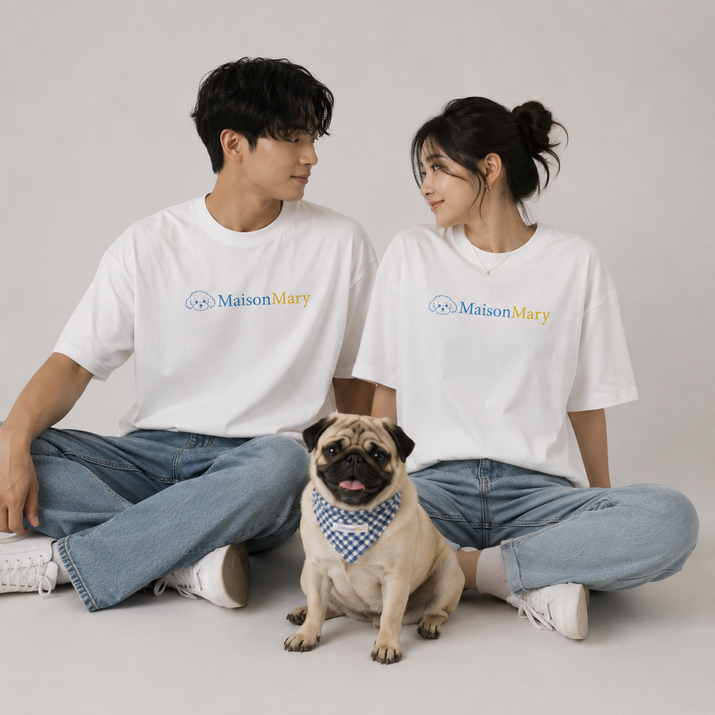 A man and a woman sitting cross-legged on a light gray studio floor, both wearing white Maison Mary graphic T-shirts and light blue jeans, with a small pug in front wearing a blue-and-white checkered bandana.