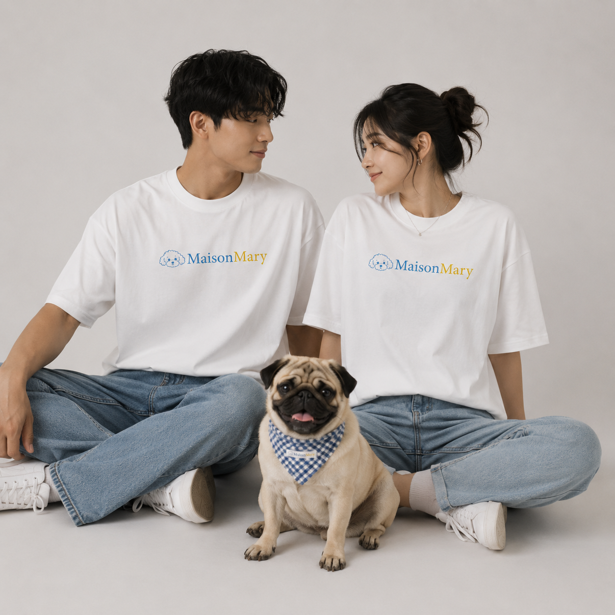 A man and a woman sitting cross-legged on a light gray studio floor, both wearing white Maison Mary graphic T-shirts and light blue jeans, with a small pug in front wearing a blue-and-white checkered bandana.